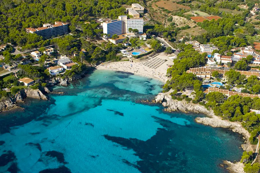 Aerial view of a sandy beach with turquoise water, surrounded by green trees and buildings.