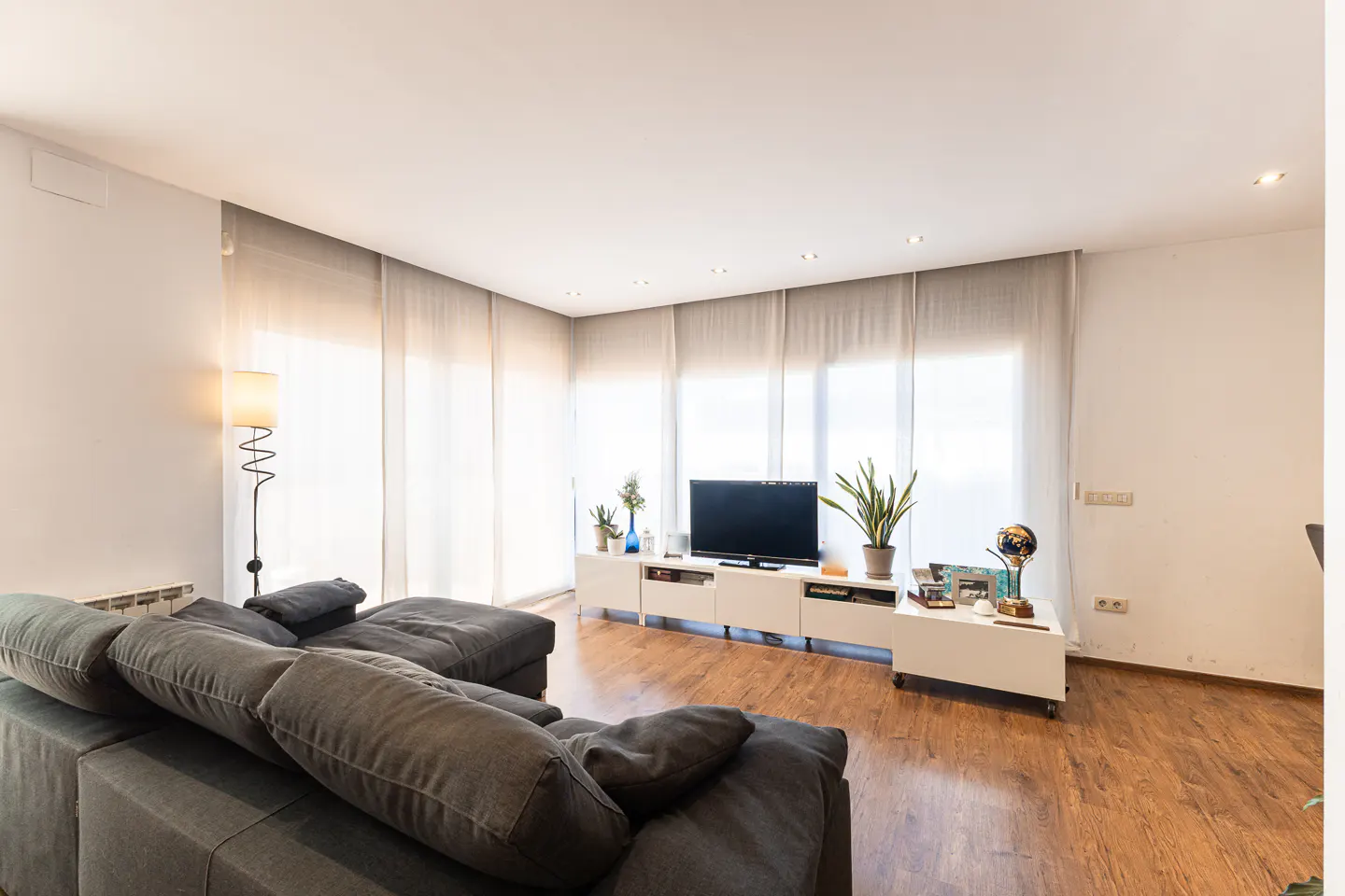Living room with a gray sectional sofa, wood floors, white walls, and a TV on a white media console. Light streams through sheer curtains.