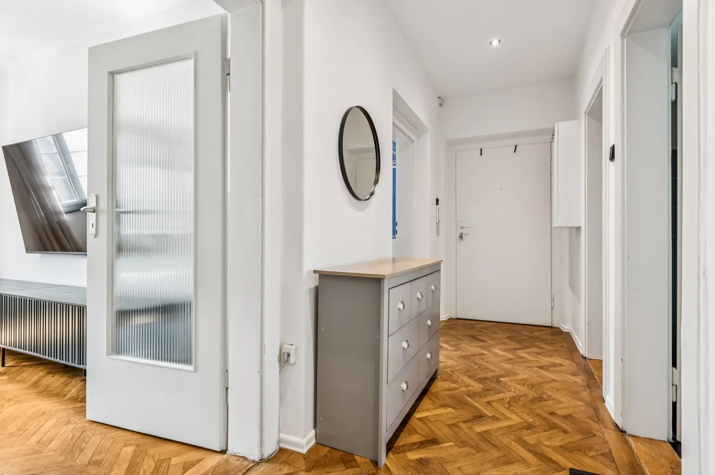 Hallway with herringbone wood floors, white walls, and a gray dresser with a round mirror above it. A glass-paneled door is on the left.