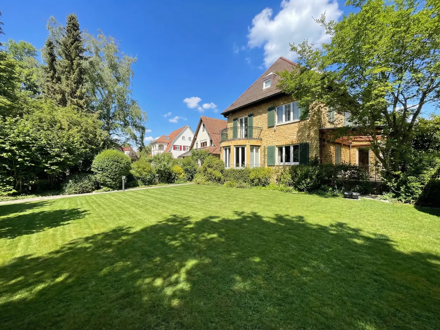 A two-story yellow stone house with green shutters and a large green lawn on a sunny day.