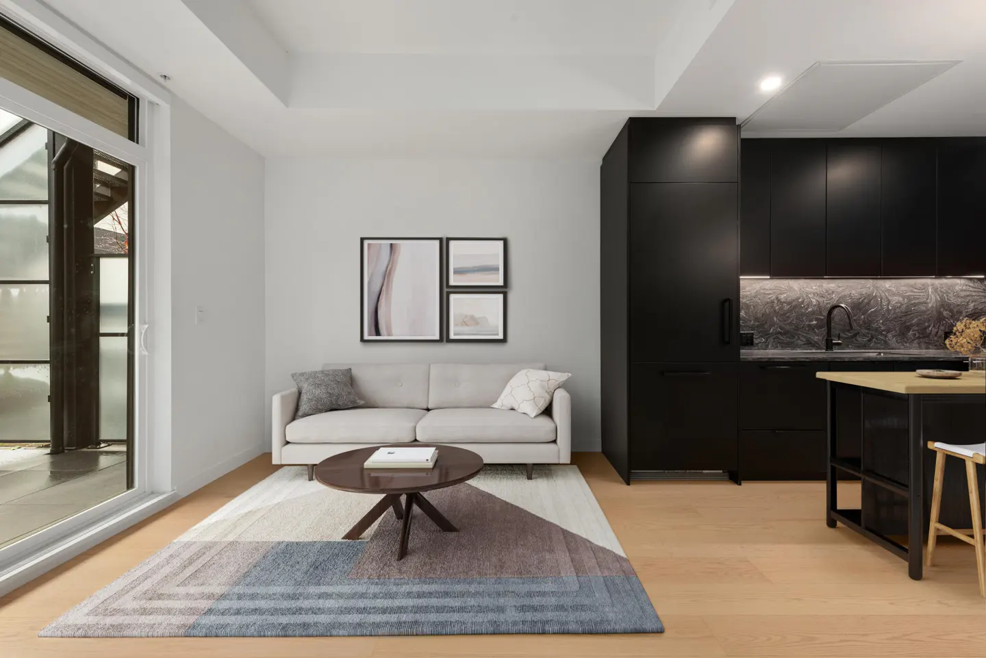 Bright living room with a gray sofa, round coffee table, and a blue and brown rug. Black kitchen cabinets are visible in the background.
