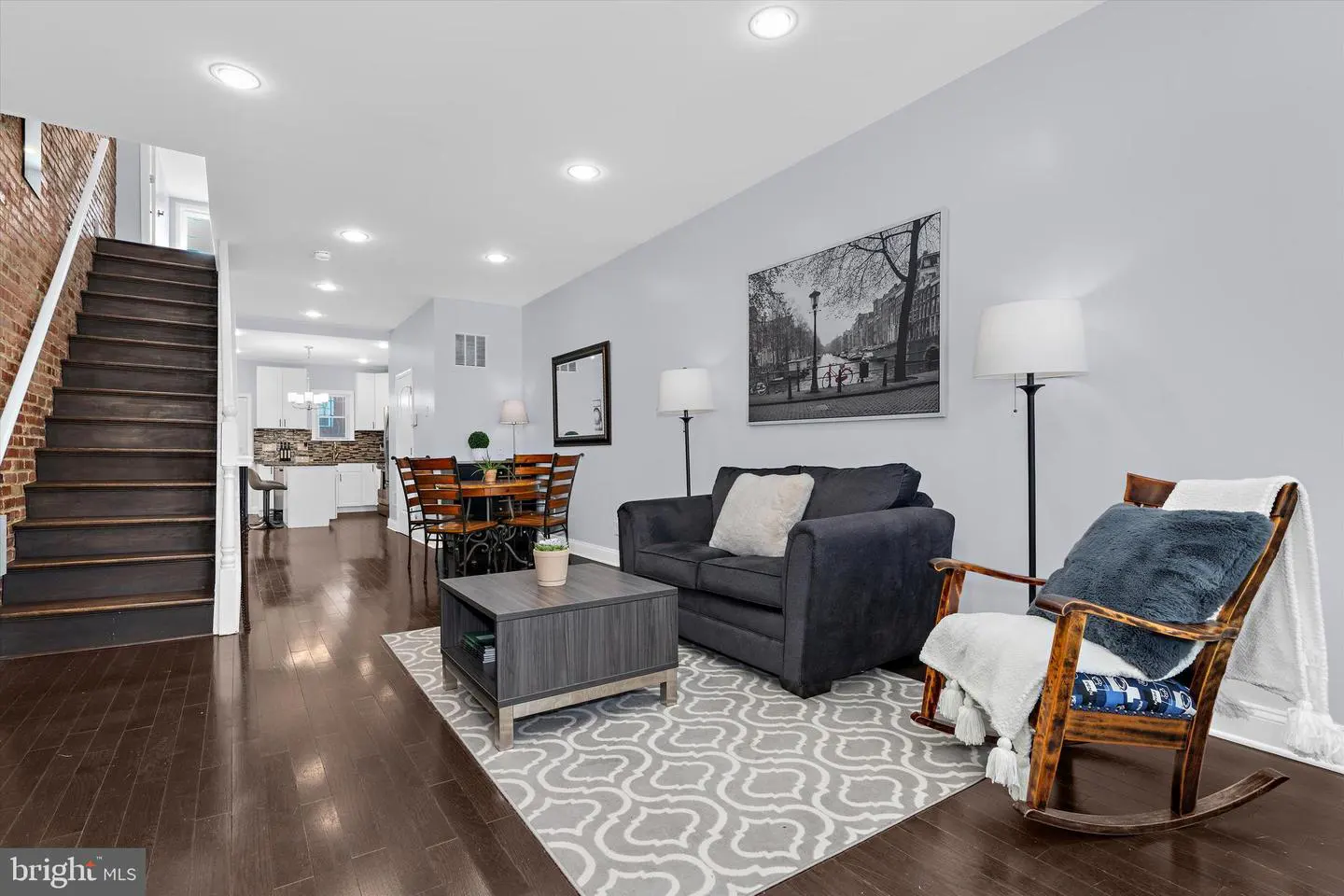 Living room with hardwood floors, gray walls, and recessed lighting. A staircase is on the left, and a kitchen is visible in the background.