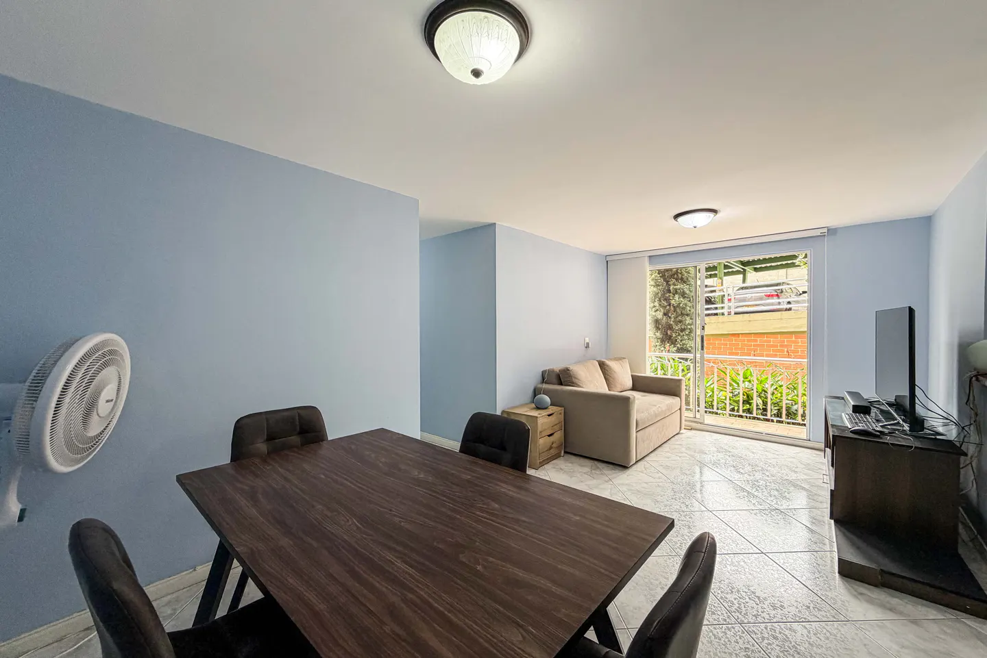 Living room with blue walls, a dark wood table with chairs, a beige sofa, and a balcony with a view of greenery.