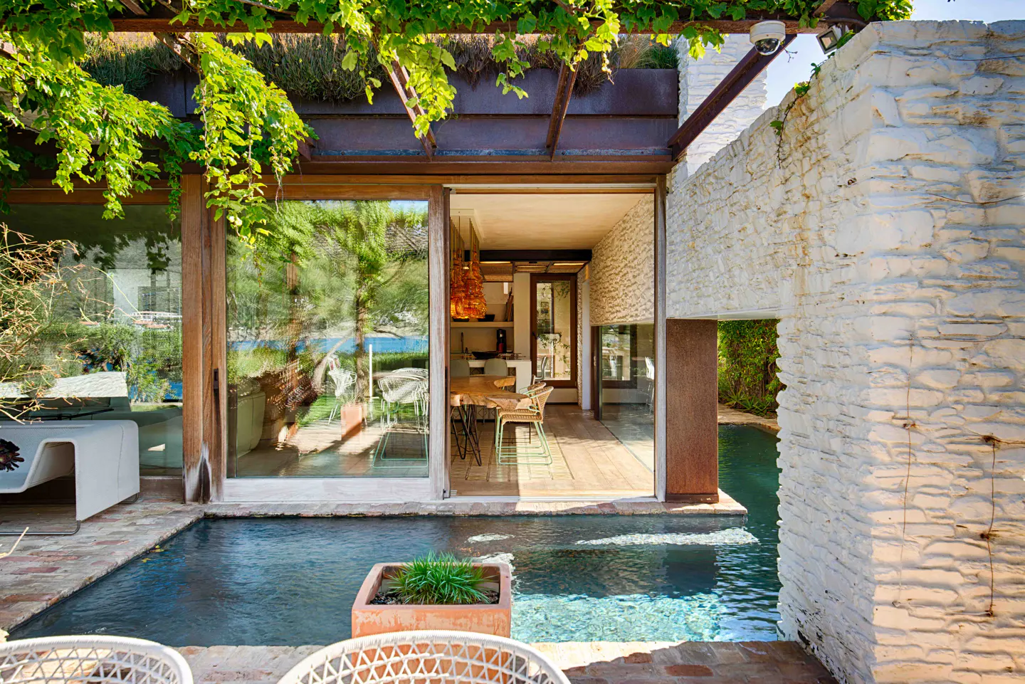 Outdoor patio with a small pool, stone wall, and open sliding glass doors to a dining area. Green vines hang from a metal pergola.