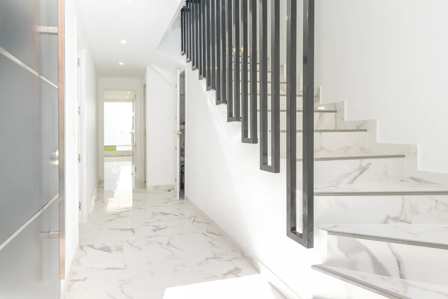 Bright hallway with marble floors and stairs. Black metal railing on the right. Doorway at the end of the hall.