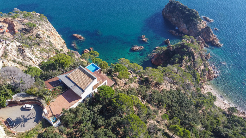 Aerial view of a modern villa with a blue pool perched on a rocky cliff overlooking the turquoise Mediterranean Sea.