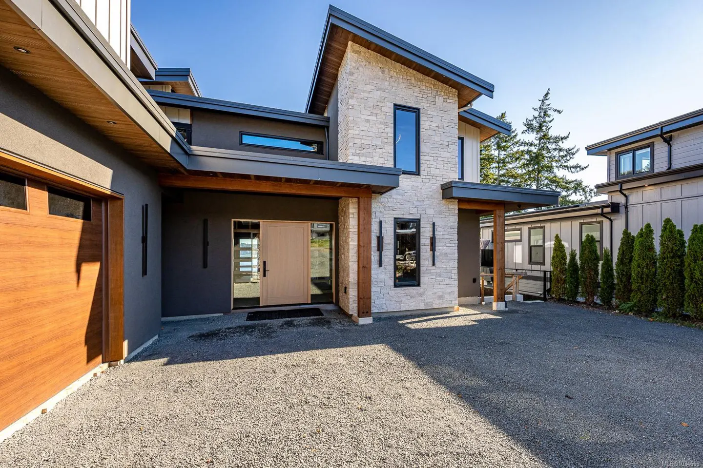 Modern home exterior with a light stone facade, dark gray accents, and a wooden garage door. A gravel driveway leads to the front entrance.