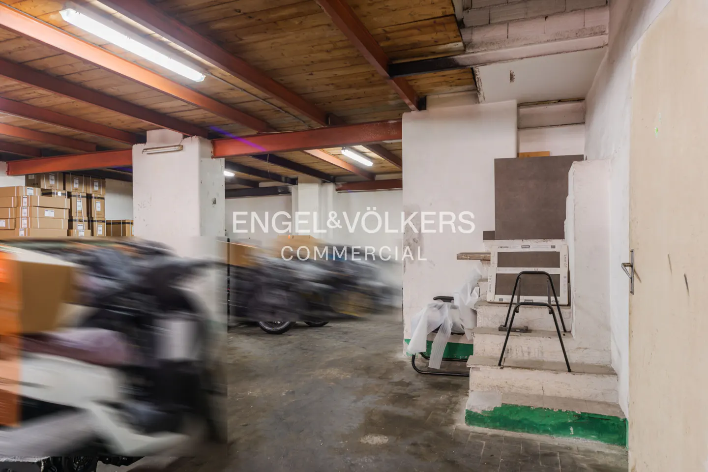 A commercial warehouse interior with a wooden ceiling, white walls, and concrete floors. Boxes and equipment are visible.