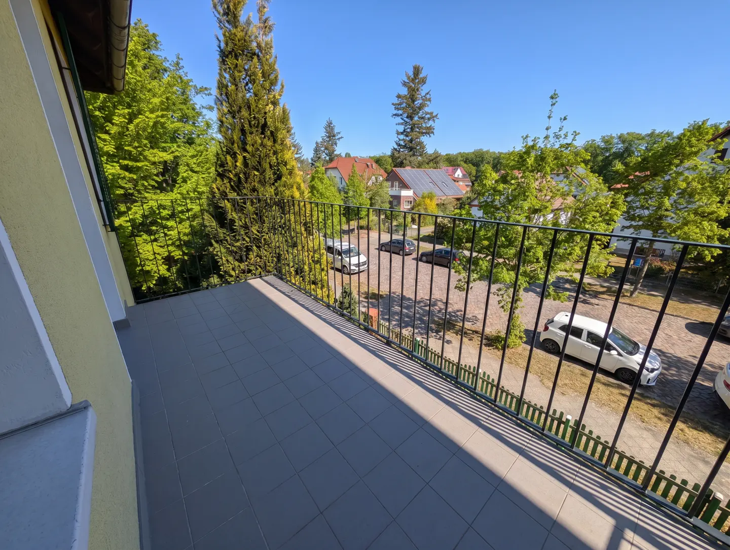 View from a balcony with gray tiles and black railings, overlooking a parking lot with cars and green trees under a blue sky.