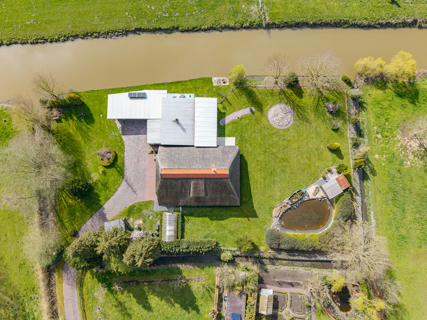 Aerial view of a thatched roof house with a pond, green lawn, and a river in the background.