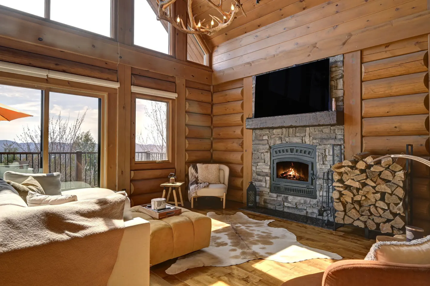 Cozy log cabin living room with stone fireplace, TV, and antler chandelier. Sofa, chair, and cowhide rug add warmth. Large windows offer scenic views.