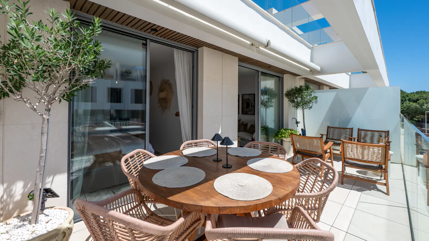 Outdoor patio with a round wooden table, wicker chairs, and place settings. Sliding glass doors lead inside. A potted tree adds greenery.