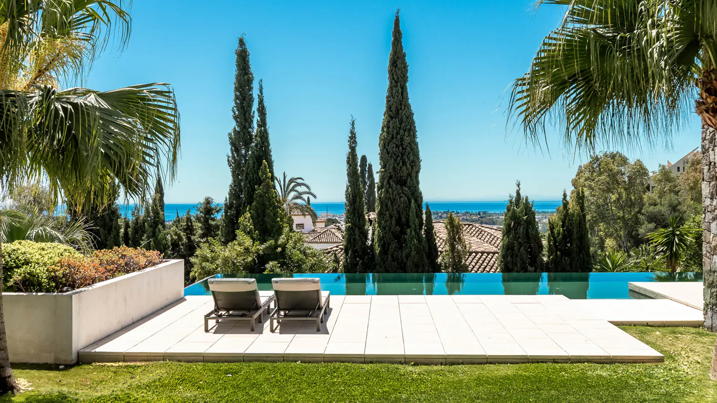 Infinity pool with two lounge chairs overlooking the ocean, surrounded by trees and a clear blue sky.