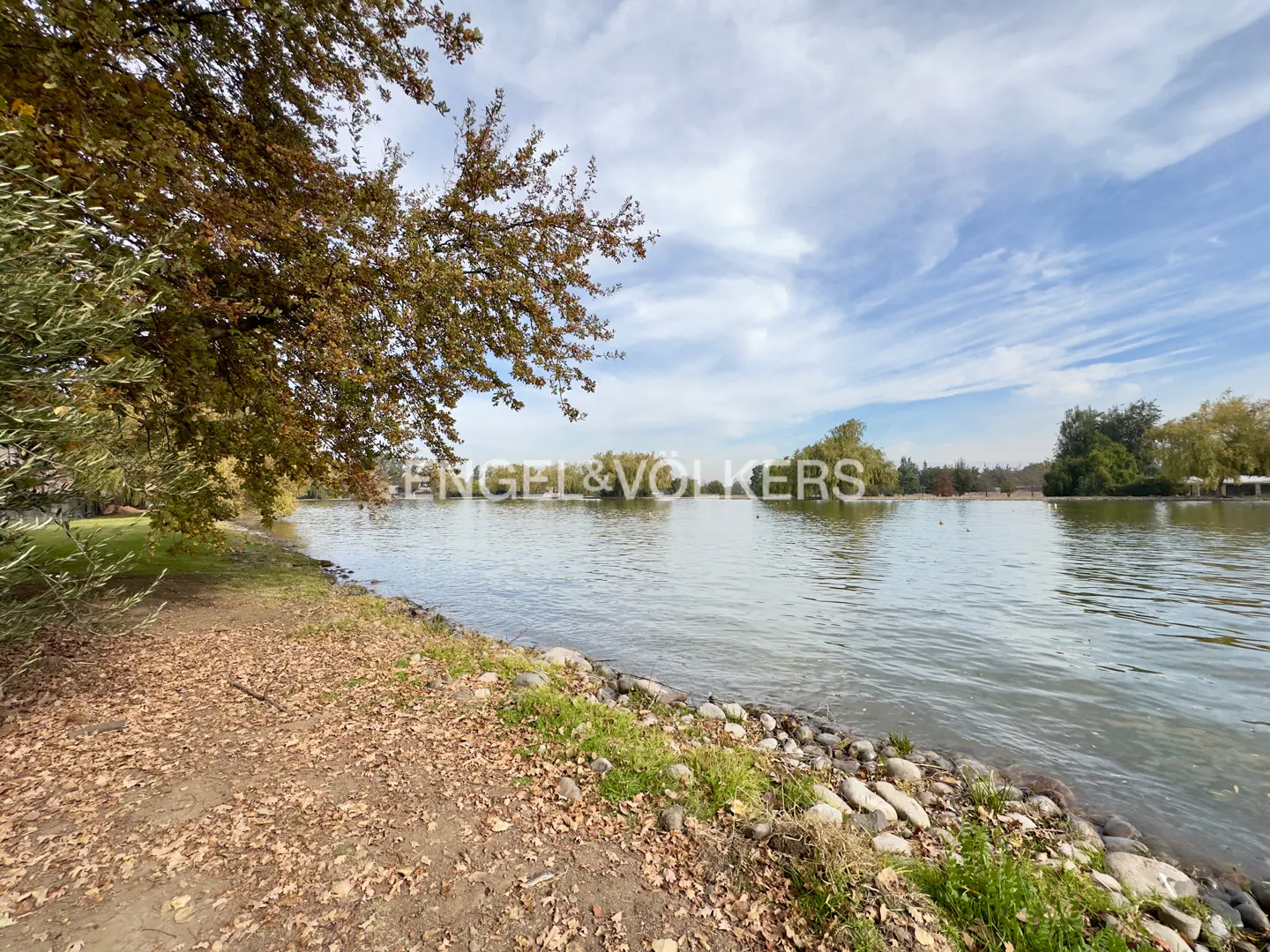 A scenic view of a calm lake with a rocky shoreline, autumn trees, and a blue sky with light clouds.