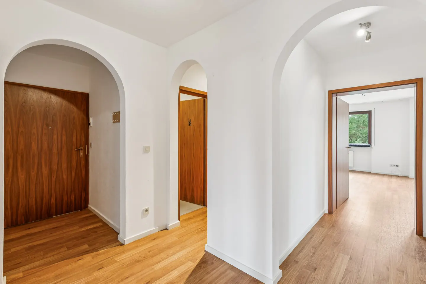 Hallway with arched doorways, white walls, and wood floors. Three wood doors are visible. One room has a window.