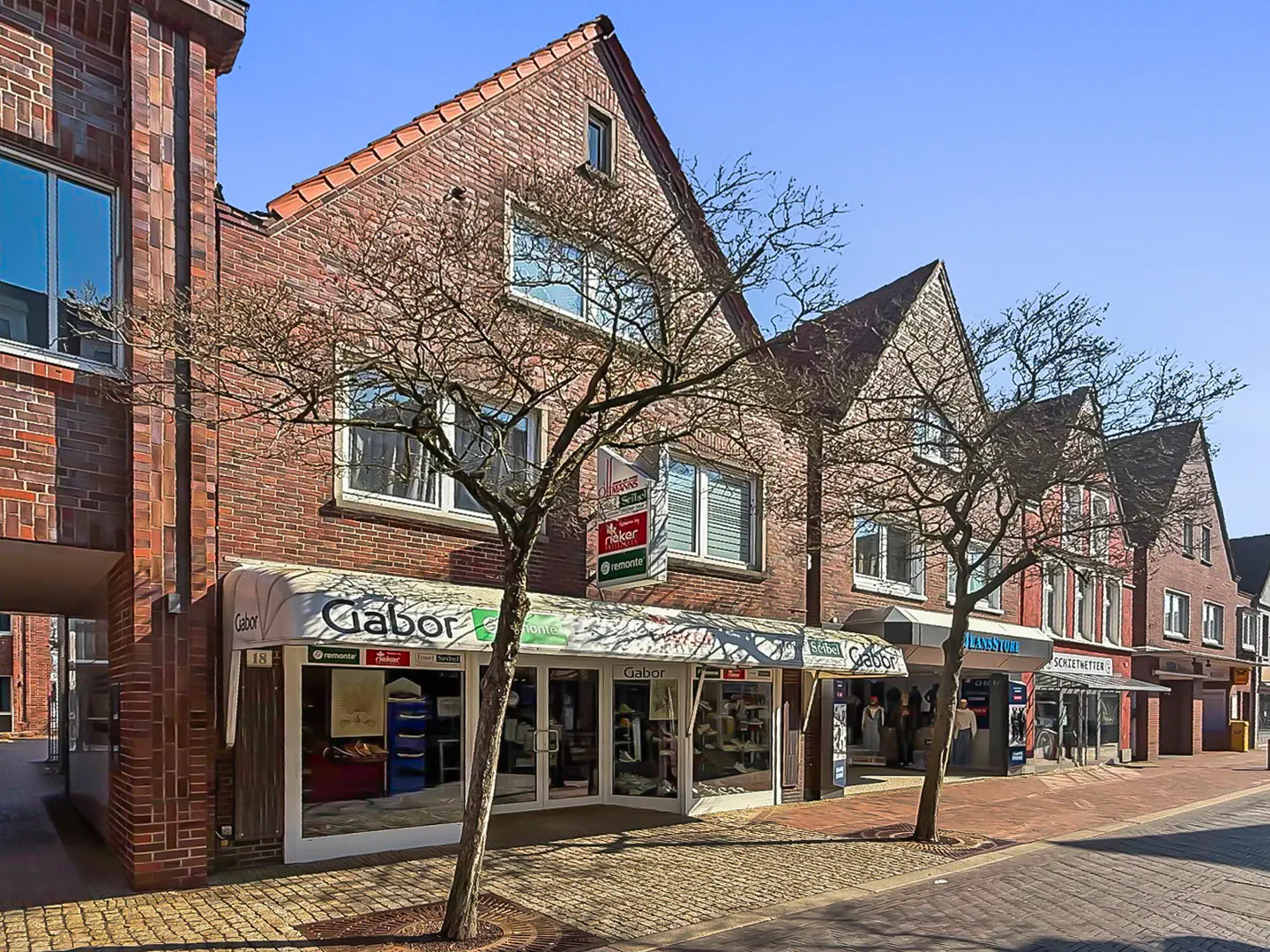 Brick buildings line a street with shops, including Gabor, under a clear blue sky. Bare trees stand in front.