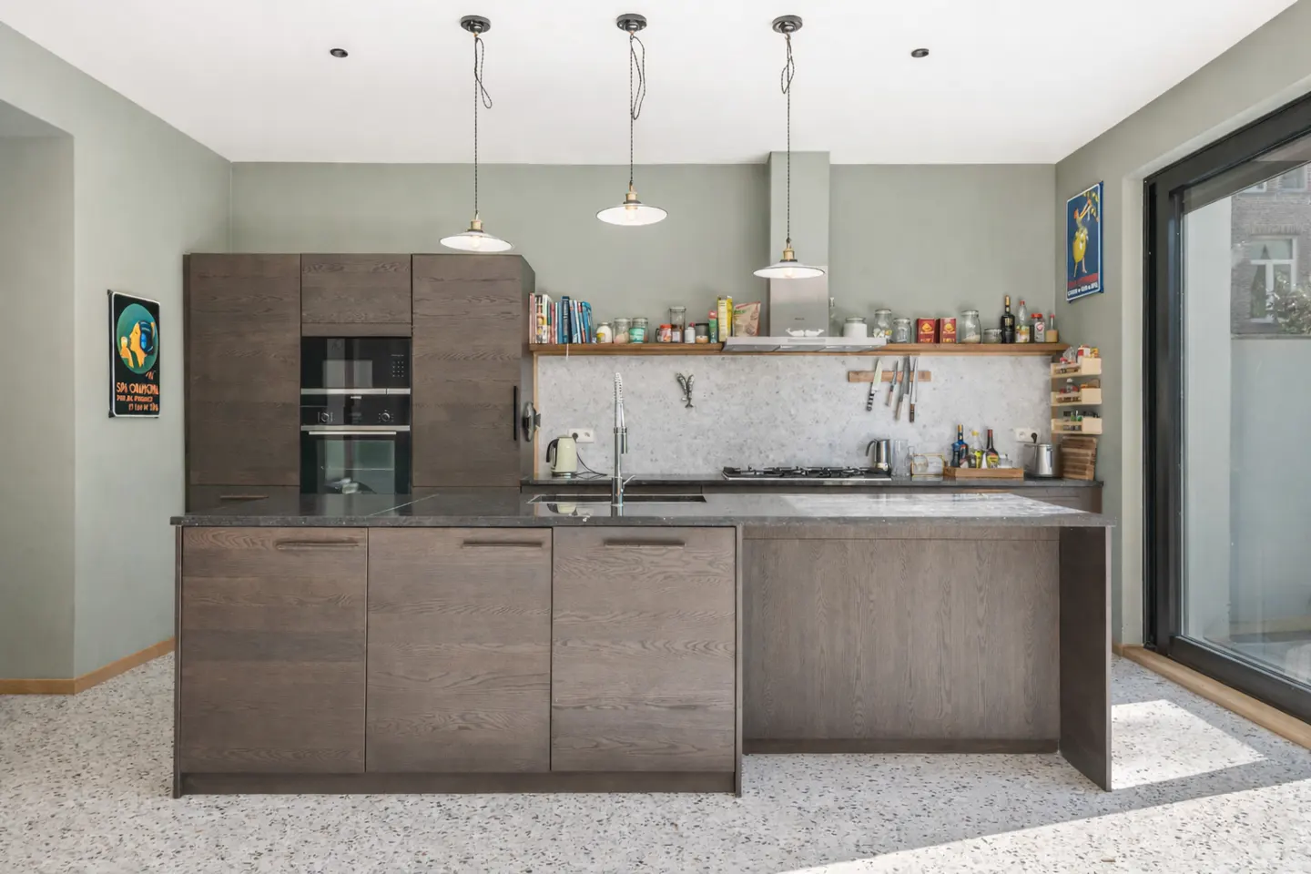 A modern kitchen with gray cabinets, a marble backsplash, and three pendant lights. A shelf holds jars and bottles. A sliding glass door leads outside.
