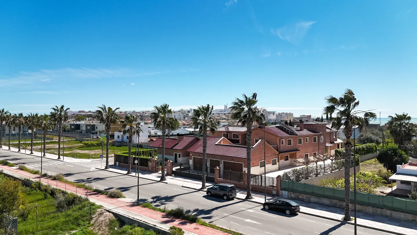 Street view of a red brick house with palm trees, cars on the road, and a clear blue sky.