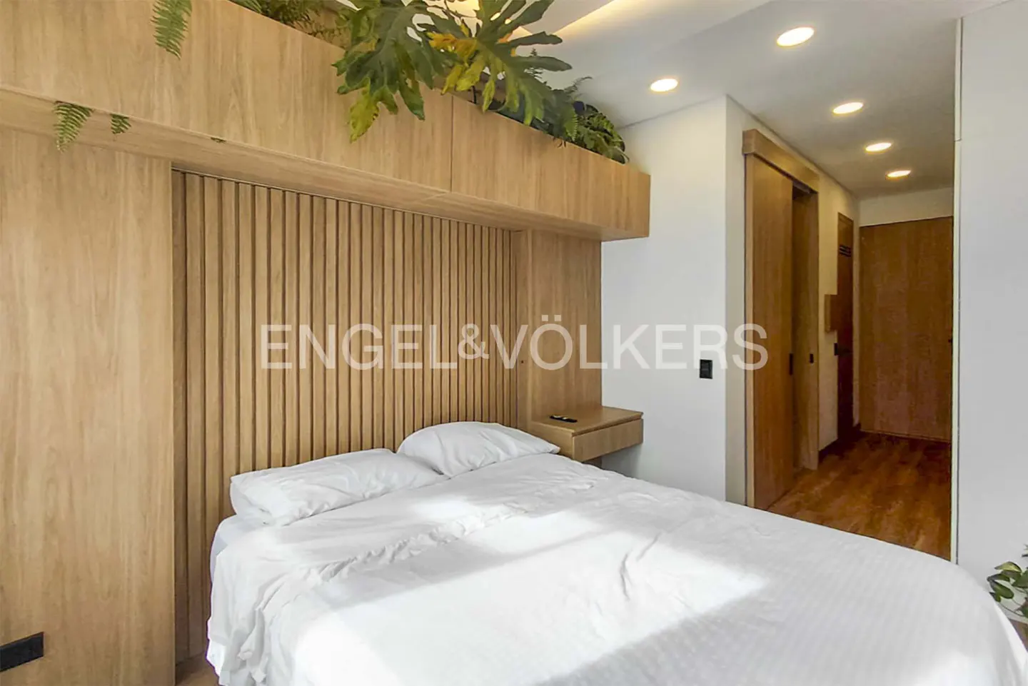 Modern bedroom with a white bed, light wood slatted headboard, overhead storage with plants, and recessed ceiling lighting.