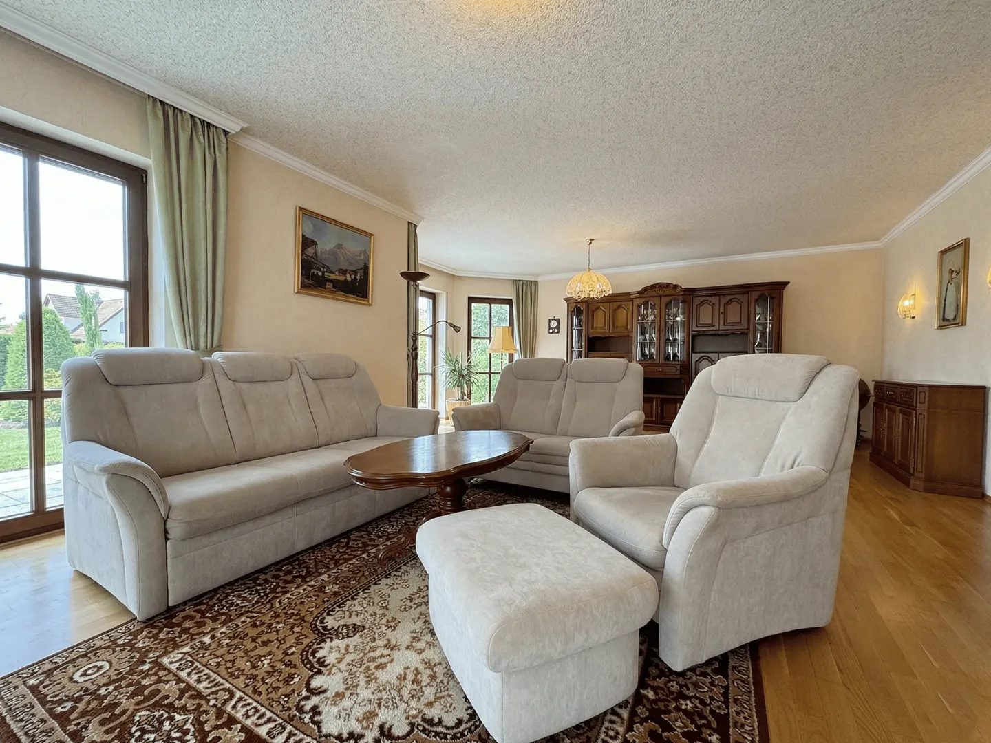 A living room with a beige sofa, armchair, ottoman, and a brown patterned rug. A wooden coffee table sits in the center.