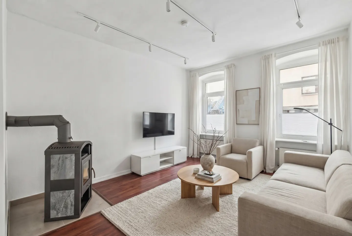 Bright living room with white walls, wood floors, and a fireplace. Beige sofa and armchair face a round coffee table and TV. Windows have sheer white curtains.