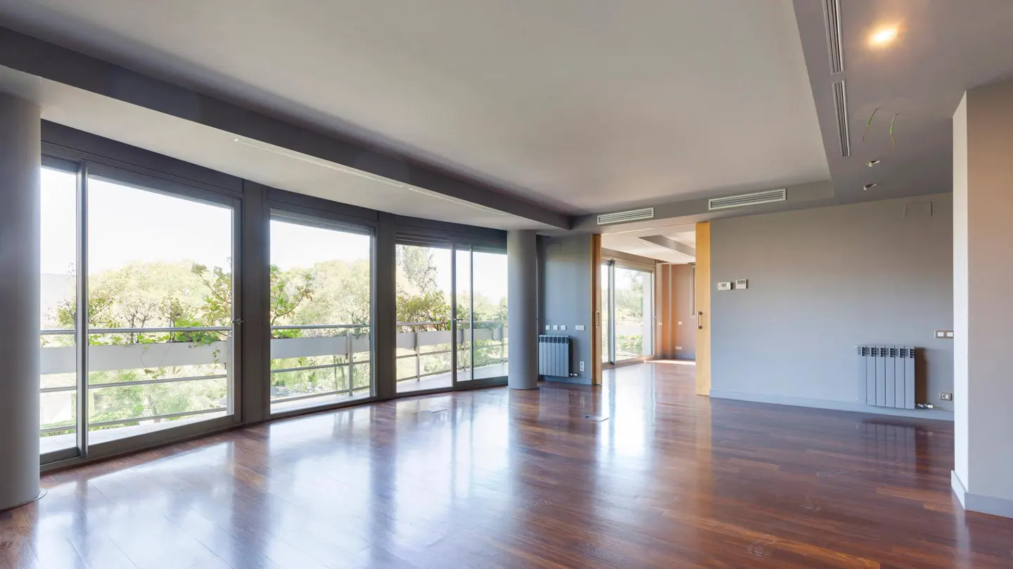 An empty room with hardwood floors, gray walls, and large windows overlooking trees and a balcony.