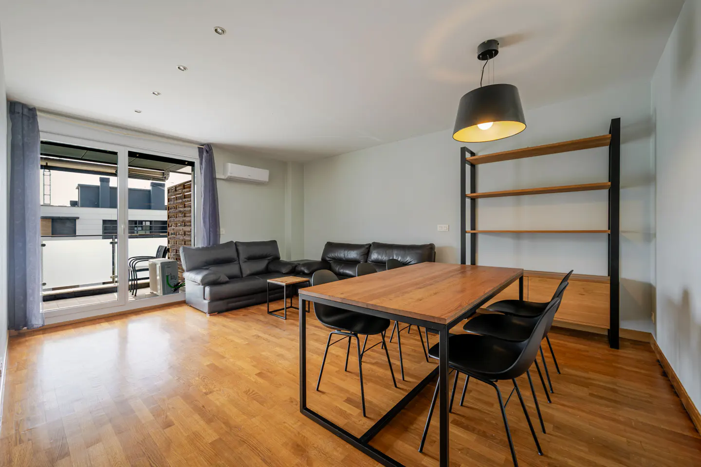 A bright living room with wood floors, a dining table with black chairs, and a black leather sofa. A balcony is visible through sliding glass doors.