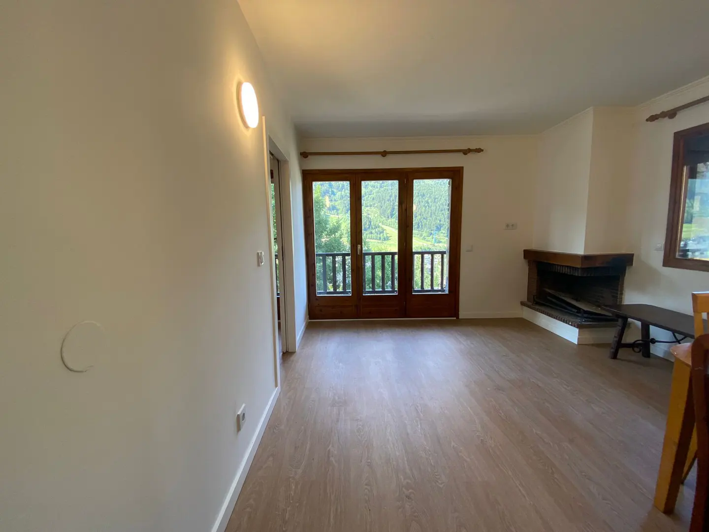 Bright living room with wood floors, white walls, and a fireplace. Balcony doors open to a view of green trees.