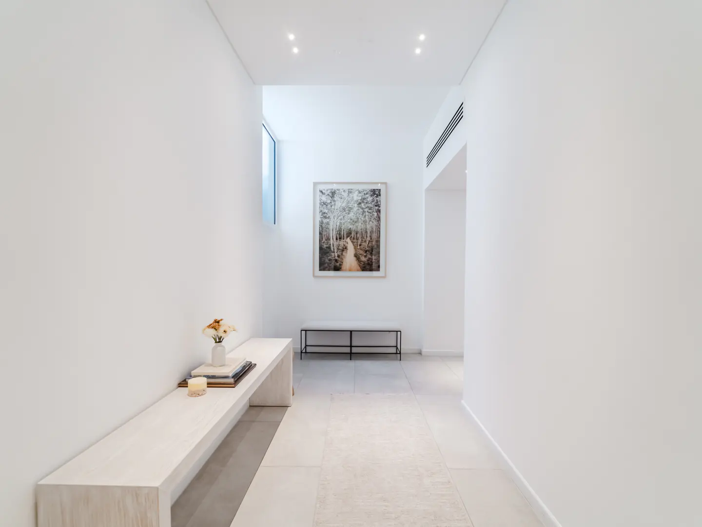 Bright, white hallway with a bench, books, flowers, and a framed forest print. A small window lets in natural light.