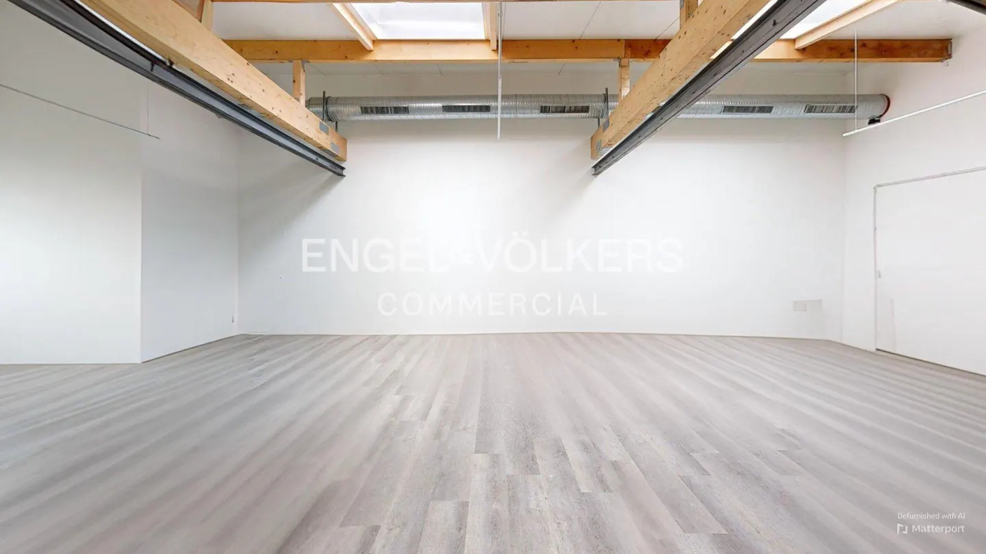 Empty commercial space with light gray wood floors, white walls, and exposed wooden beams. "Engel & Völkers Commercial" is printed on the wall.