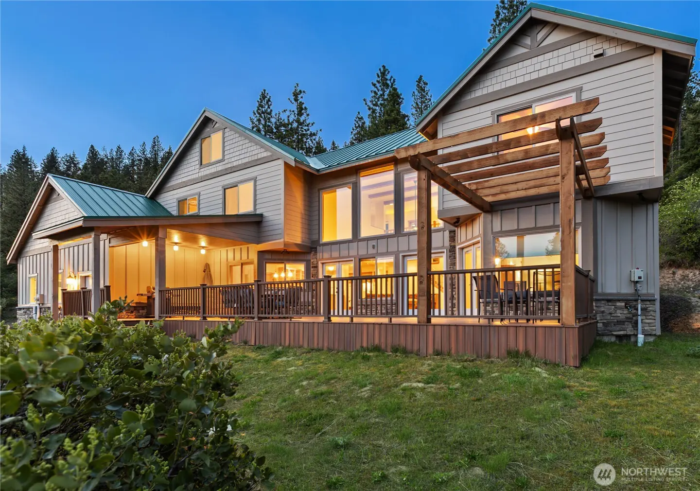 Large two-story home with a green metal roof, wood deck, and pergola, set against a forest at dusk.