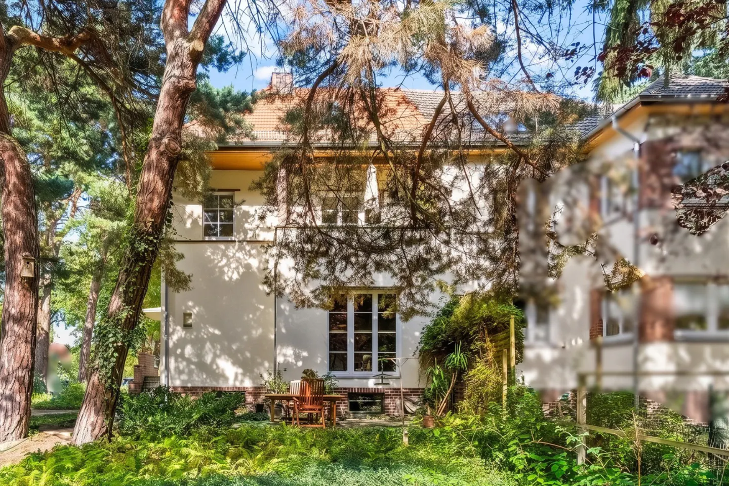 Exterior view of a two-story white house with a red tile roof, surrounded by lush greenery and trees. A wooden table and chairs sit on the patio.