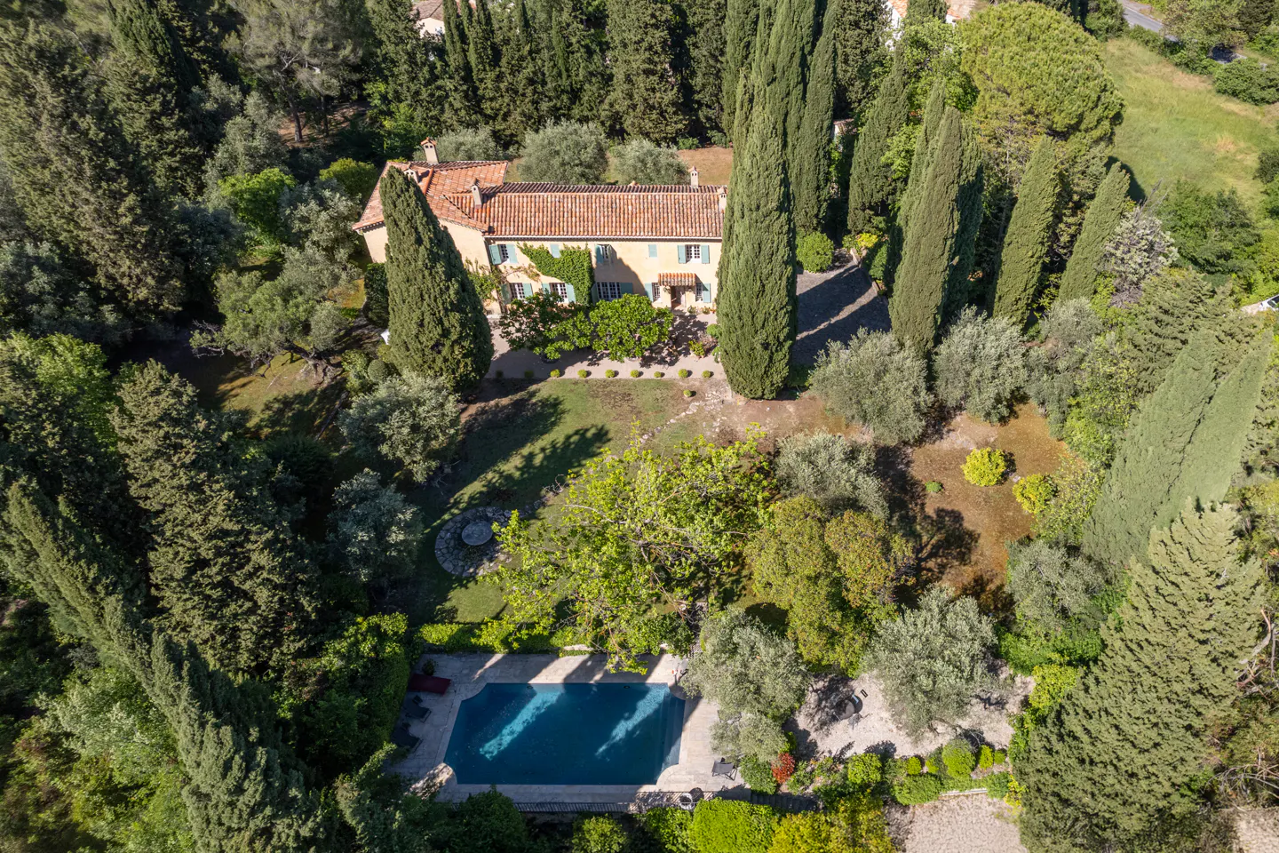 Aerial view of a yellow villa with a red tile roof, surrounded by tall green trees and a blue swimming pool.