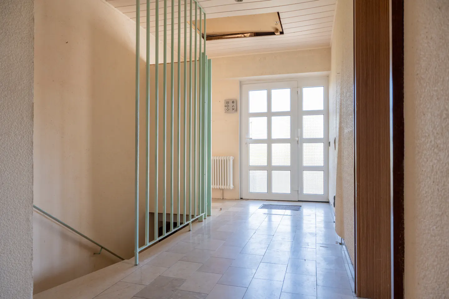 Hallway with stairs, light-green railing, and white double doors. Tiled floor and beige walls. Radiator visible.