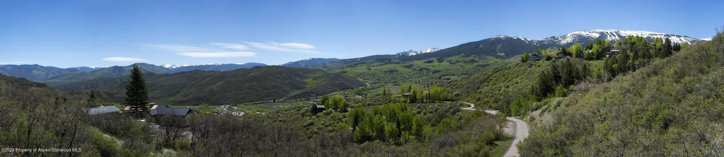 Panoramic view of a lush green valley with snow-capped mountains in the background under a clear blue sky. A winding road cuts through the landscape.