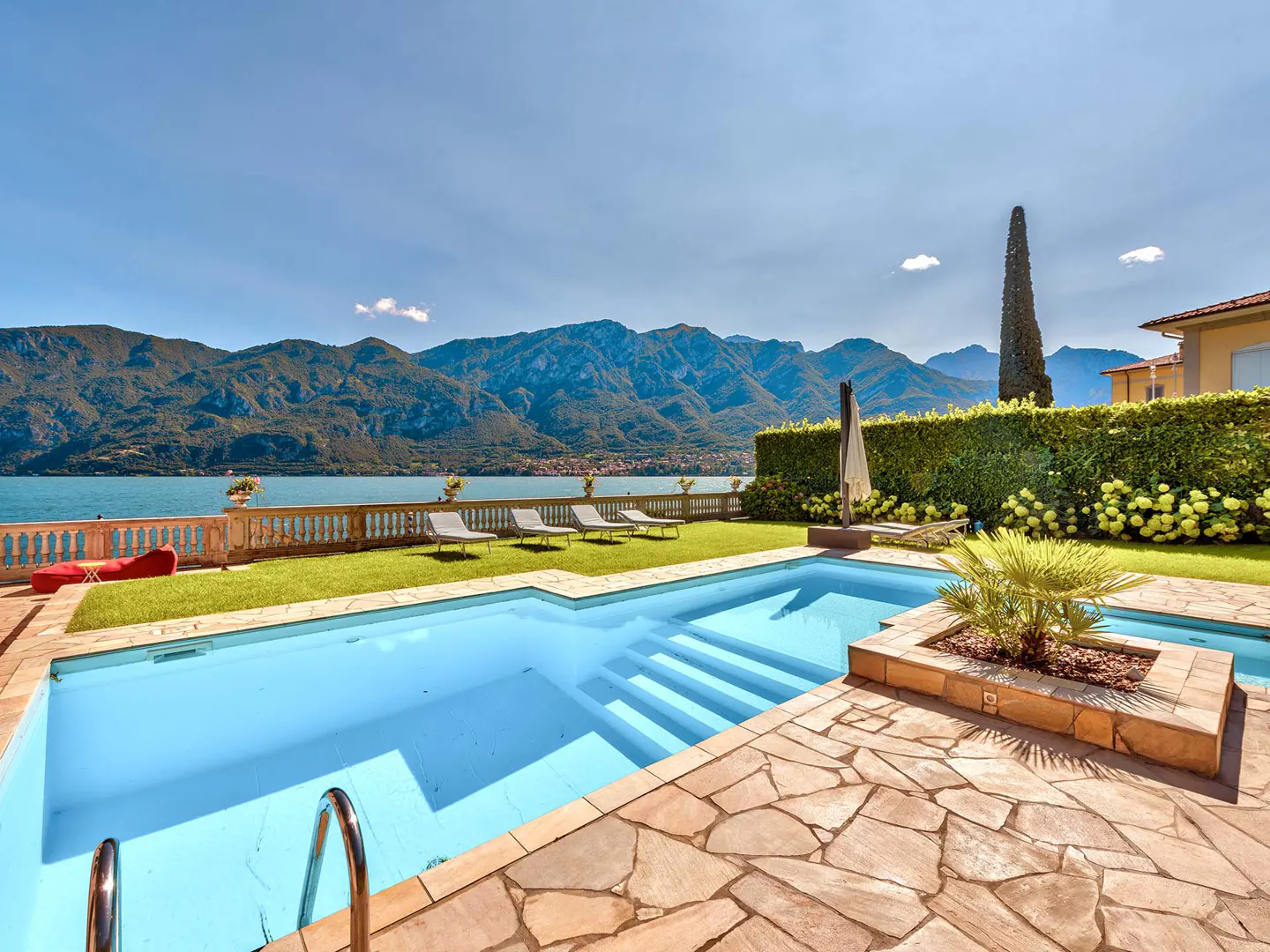 A blue swimming pool with stone patio, green grass, and lounge chairs overlooks a lake and mountains under a blue sky.