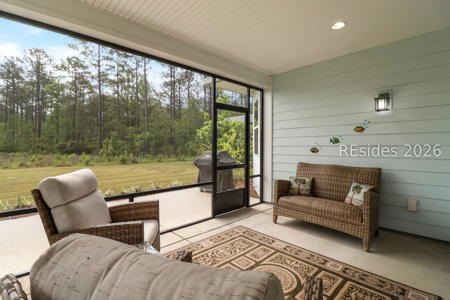Screened porch with wicker furniture, including a sofa and chair, on a patterned rug. A grill is visible outside, with trees in the background.