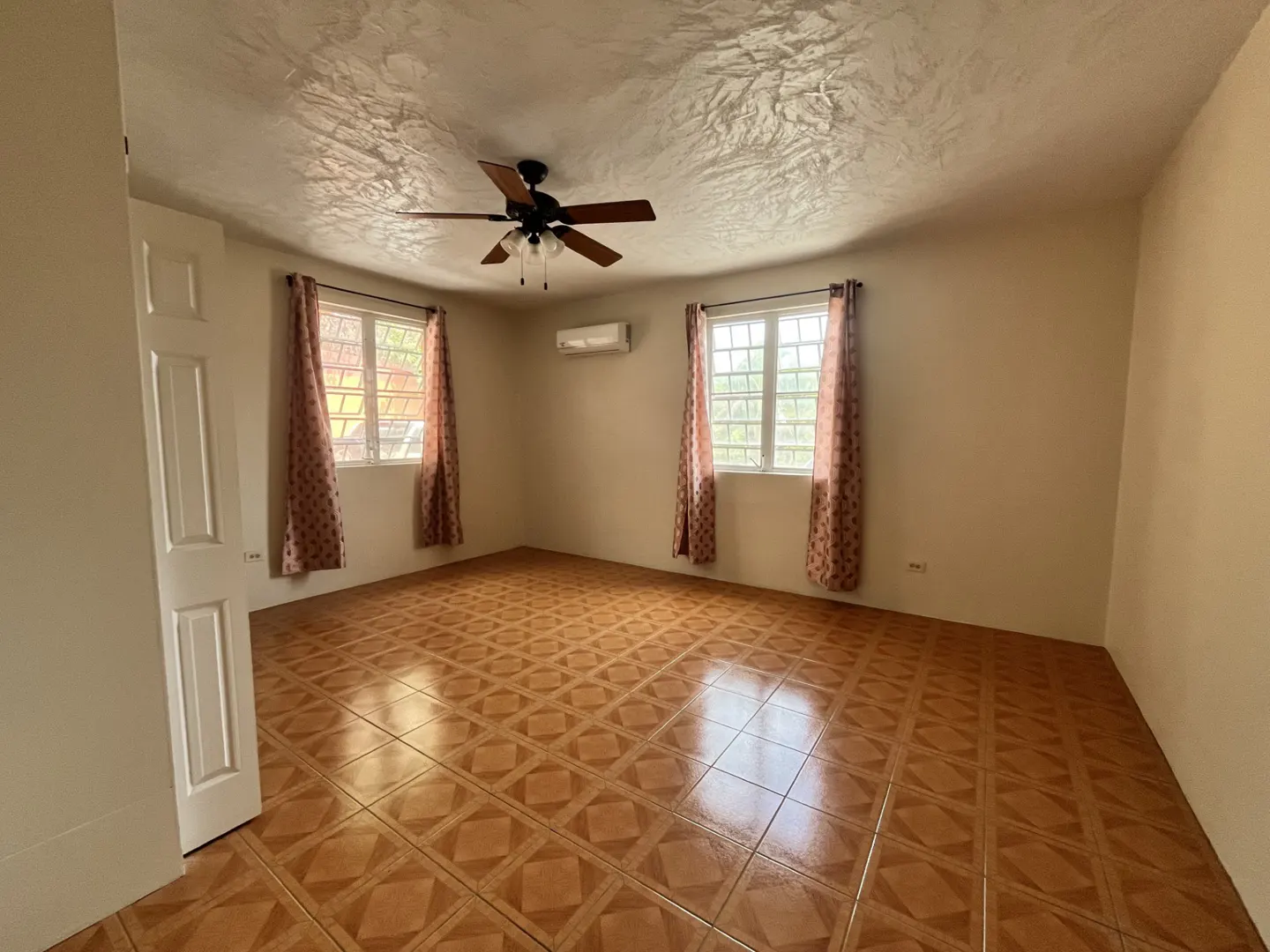 Empty room with beige walls, patterned tile floors, two windows with curtains, a ceiling fan, and a wall-mounted AC unit.