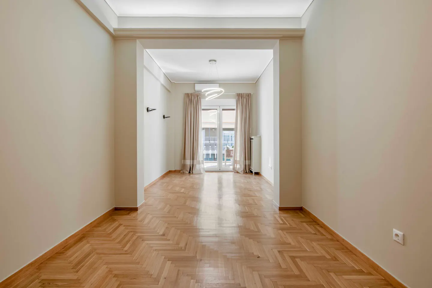 Empty room with herringbone wood floor, beige walls, and a doorway to a balcony with curtains.