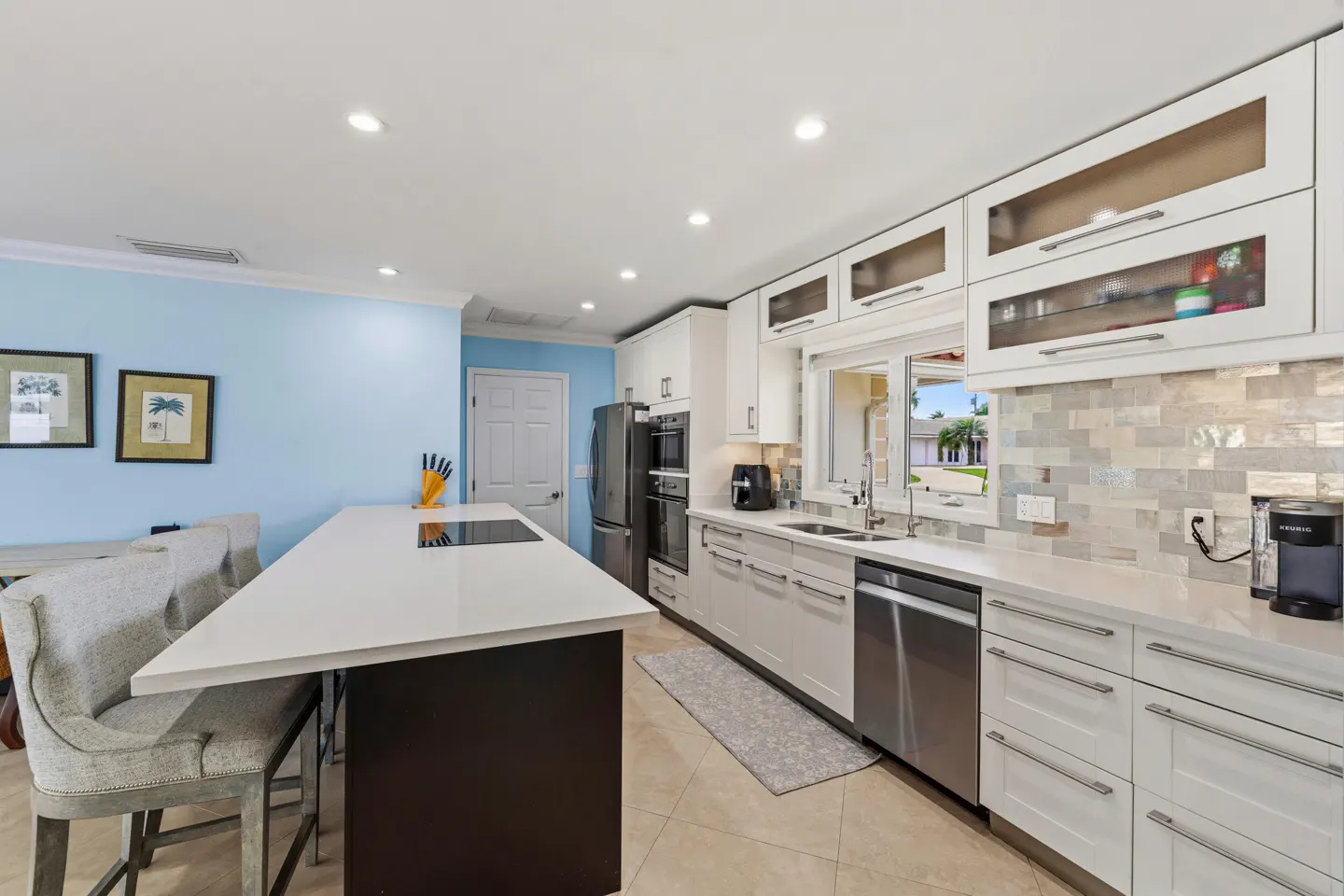 A modern kitchen with a white countertop island, gray chairs, and light blue walls. Stainless steel appliances and white cabinets are visible.