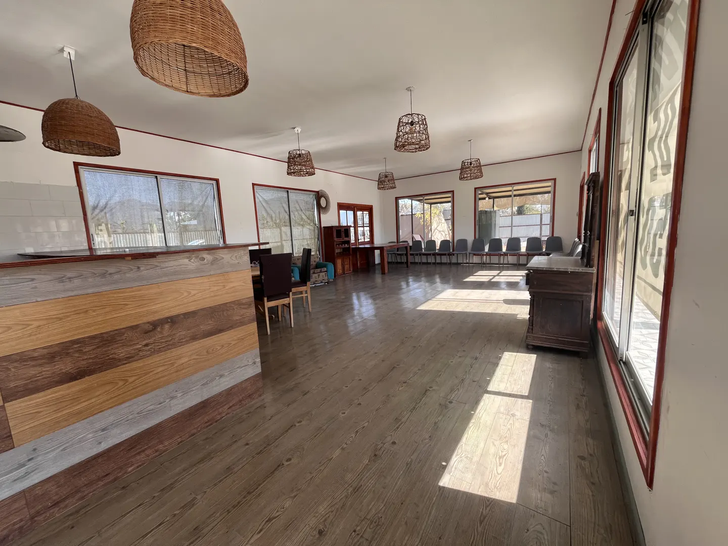 A bright, empty room with wood floors, white walls, and brown trim. Wicker light fixtures hang from the ceiling. Windows line the walls.