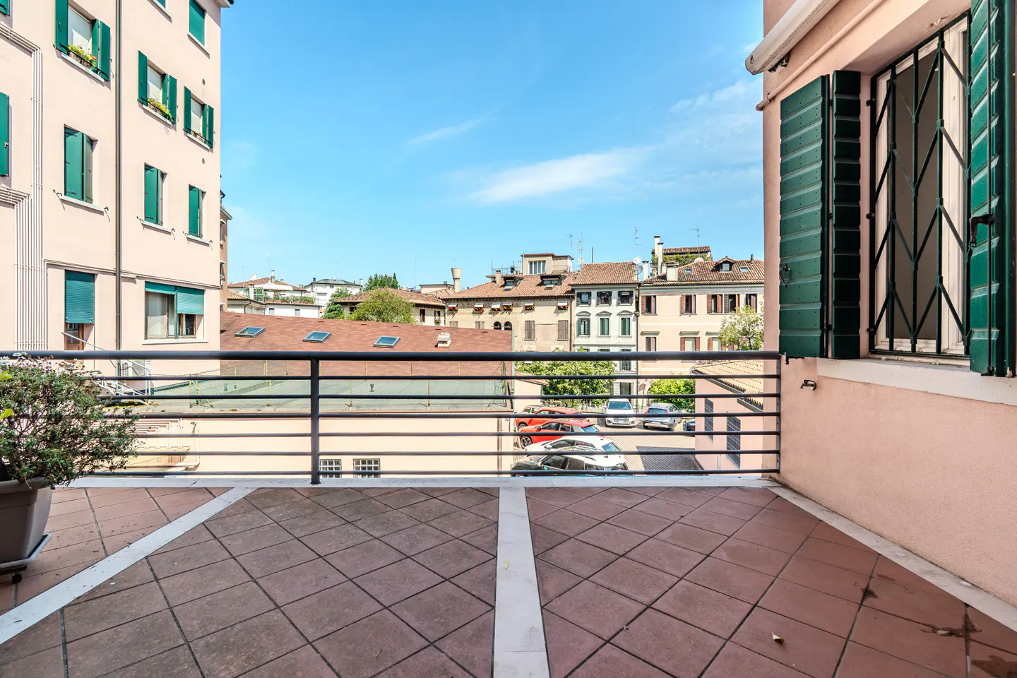 Tiled balcony with metal railing overlooking a European city street with historic buildings and parked cars under a blue sky.