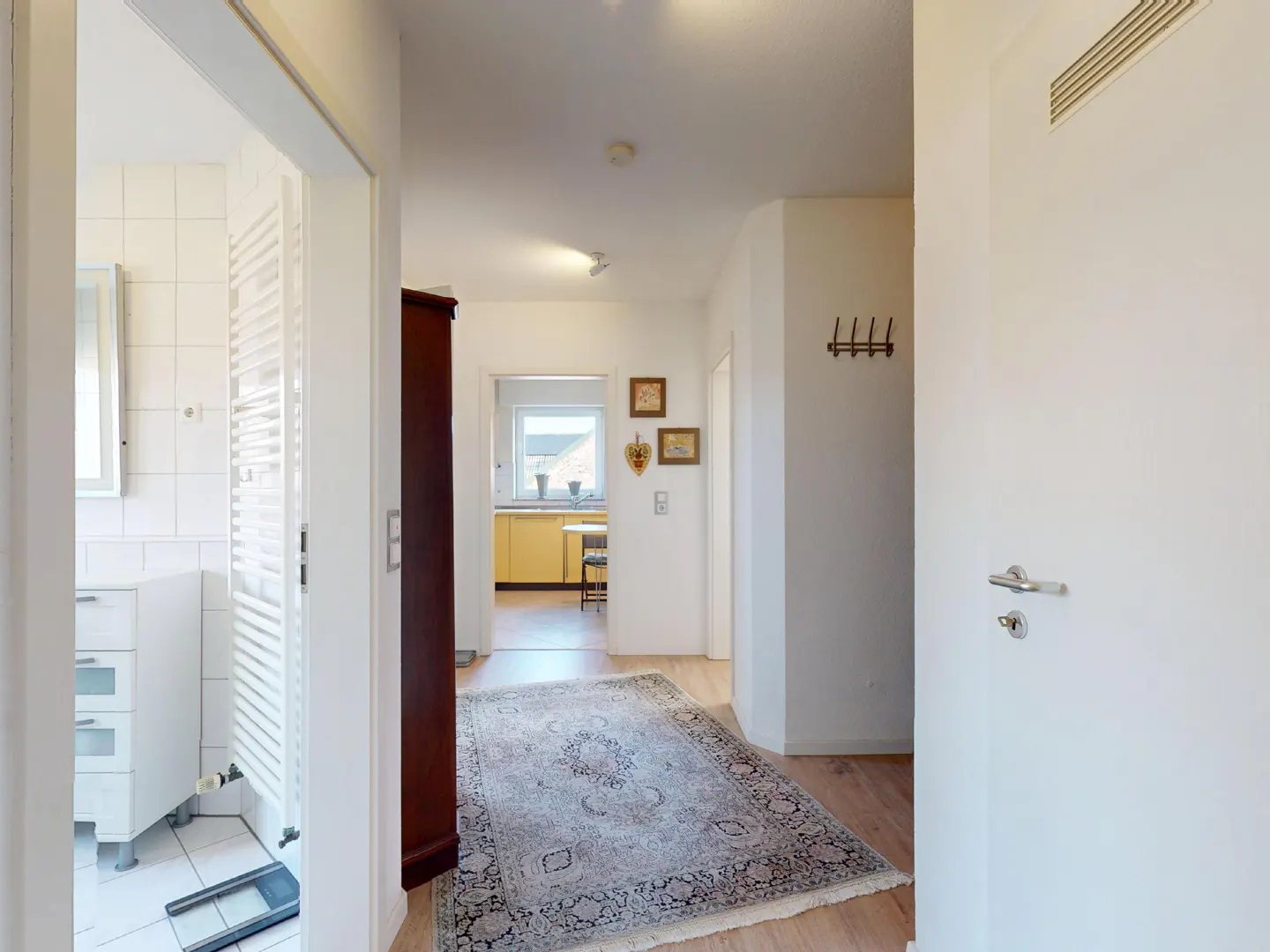 Hallway view with a patterned rug on a wood floor. A bathroom is to the left, and a kitchen is visible in the background.