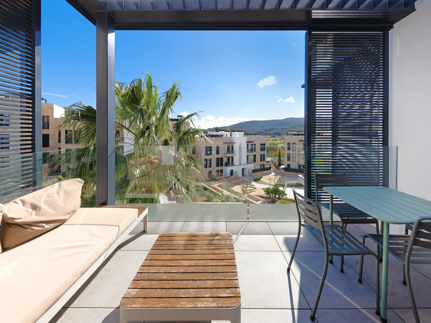 Balcony with a beige lounge chair, wooden table, and green table with chairs. Palm trees and buildings are visible in the background.