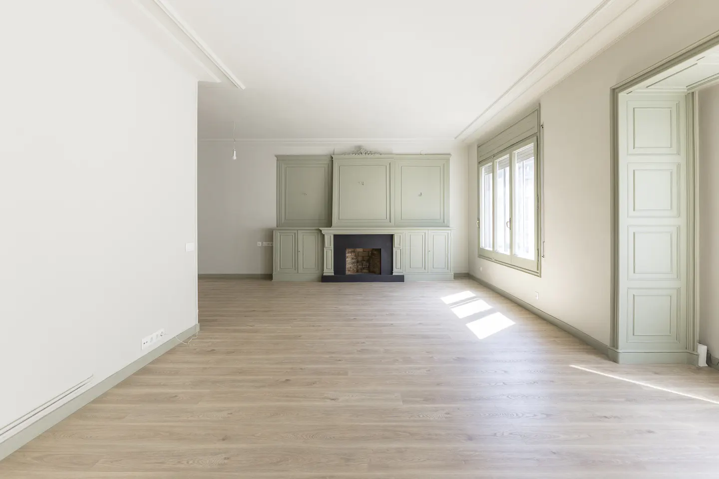 Bright, empty room with light wood floors, a sage green fireplace and built-in cabinets, and a window with sheer white curtains.