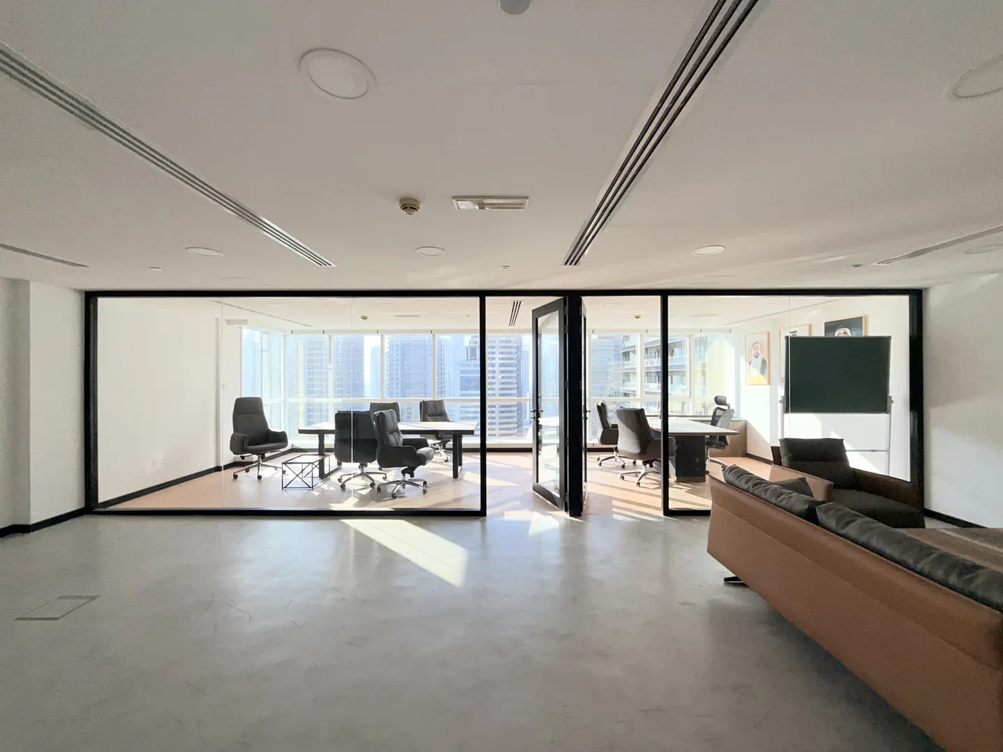 Bright, modern office space with glass-walled conference rooms, black chairs, and a brown leather sofa. Cityscape visible through windows.