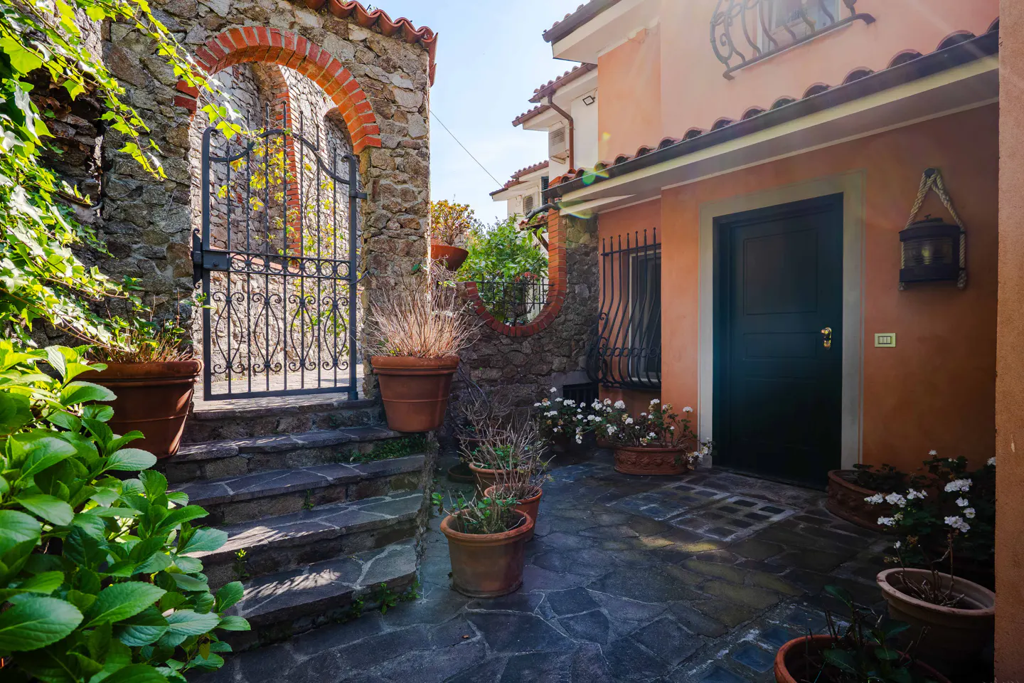 Stone courtyard with potted plants, a black iron gate in a brick archway, and a peach-colored building with a dark green door.
