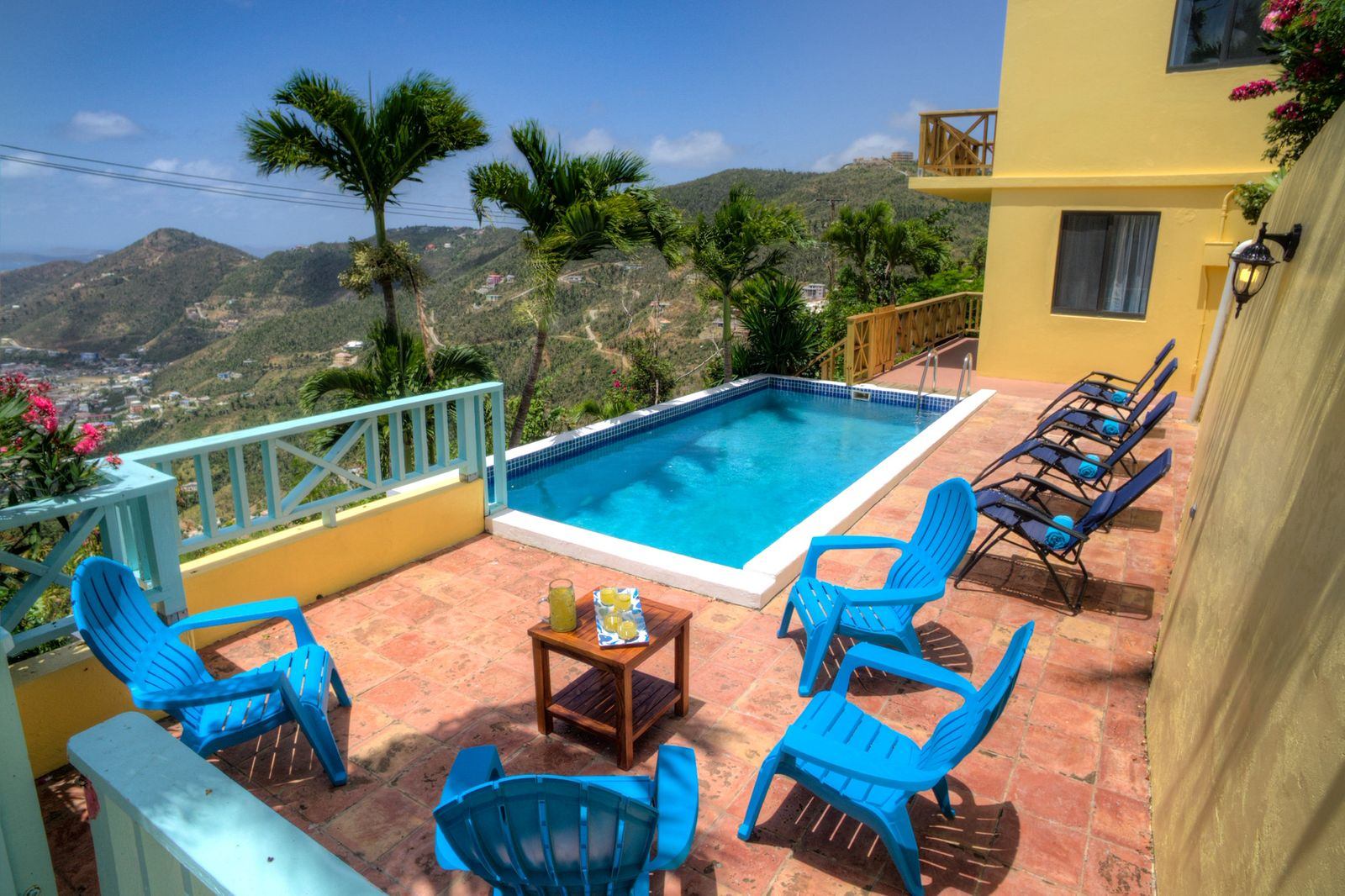 Outdoor pool area with blue chairs, a small table with drinks, and lounge chairs. Palm trees and a mountain view are in the background.