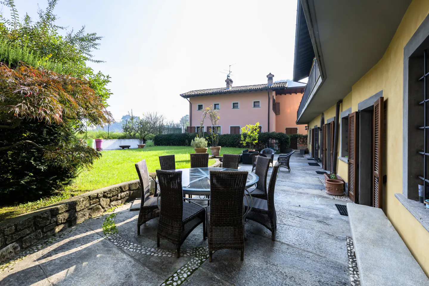 Outdoor patio with a round glass table and eight wicker chairs. A green lawn and a pink house are in the background.