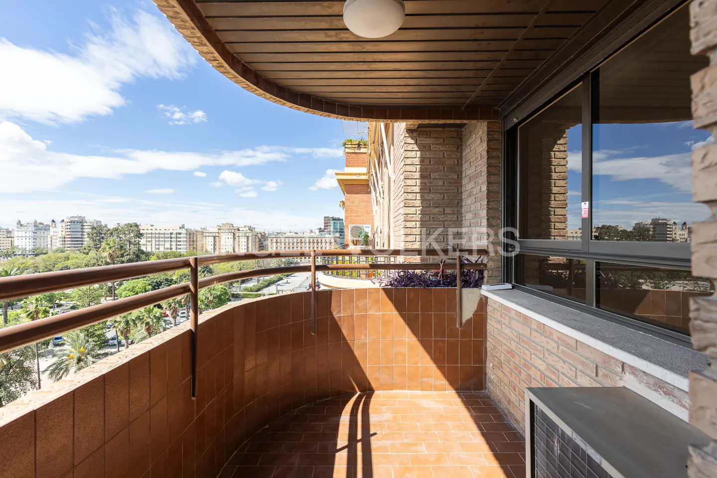A curved balcony with brown tile flooring and a metal railing overlooks a city skyline under a blue sky.